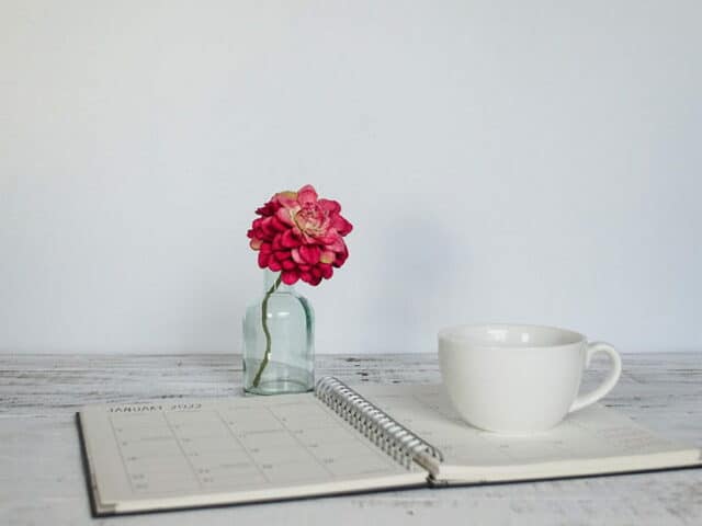 Agenda papier sur un bureau, sur lequel est posée une tasse. Un soliflore avec une fleur rose se tient à l'arrière-plan.